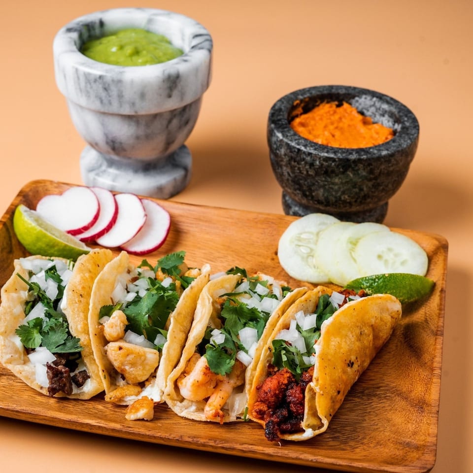 Four La Familia tacos with various fillings, cilantro, onions, lime, radishes, and sauces on a wooden tray.