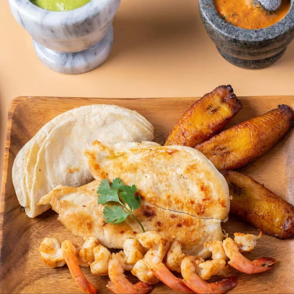 Grilled chicken and shrimp with plantains and tortillas on a wooden plate, with two molcajetes.